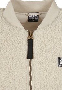 Kurtka bombowca z polarowego materiału w kolorze kremowym, z teksturowanym korpusem, żeberkowym kołnierzem, złotym zamkiem błyskawicznym oraz czarną naszywką z logo po lewej stronie.