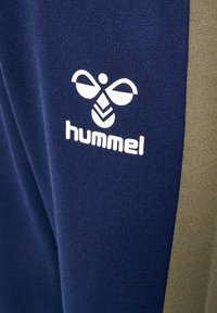 Marineblaue Jogginghosen mit olivgrünem Seitenstreifen, versehen mit einem weißen Drucklogo einer stilisierten Biene und dem Markennamen "hummel." Weicher Stoff.