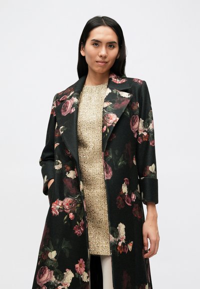 Lillemustriga, tumedat värvi mantel pikkade varrukatega, kraega ja küljetaskutega, kantud tekstuurse kuldse topi peal.