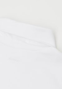 Bílý bavlněný svetr s překlopeným límcem, hladkou texturou a minimalistickým designem s jemným logem v oblasti krku.