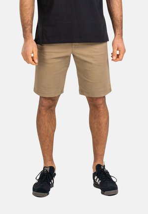 Hombre de pie vestido con pantalones cortos beige hasta la rodilla, zapatillas negras Adidas con rayas blancas y una camiseta negra, frente a un fondo liso.