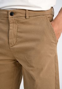 Pantalons marron à motifs en tissu texturé avec fermeture à bouton et poches latérales, présentant un motif à carreaux sur le matériau.