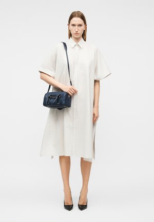 Γυναίκα που φοράει ένα φαρδύ λευκό φόρεμα με κουμπιά, κρατώντας μια μικρή χειραποσκευή σε σκούρο μπλε και μαύρες ψηλοτάκουνες γόβες, σε λευκό φόντο.