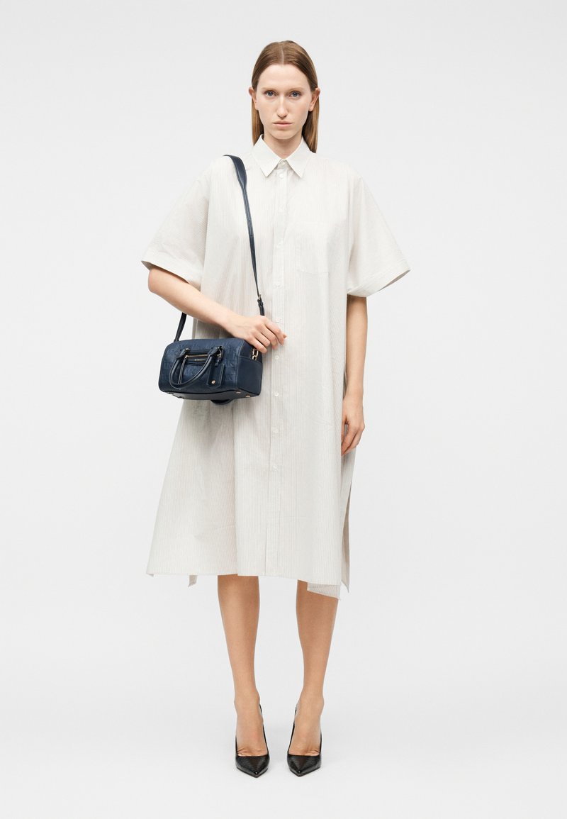 Γυναίκα που φοράει ένα φαρδύ λευκό φόρεμα με κουμπιά, κρατώντας μια μικρή χειραποσκευή σε σκούρο μπλε και μαύρες ψηλοτάκουνες γόβες, σε λευκό φόντο.