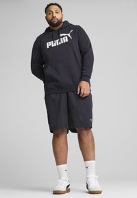 Sweat à capuche bleu marine avec le logo "PUMA" blanc, associé à un short assorti. Comprend une poche avant, des poignets côtelés et des chaussures de sport blanches avec des accents.