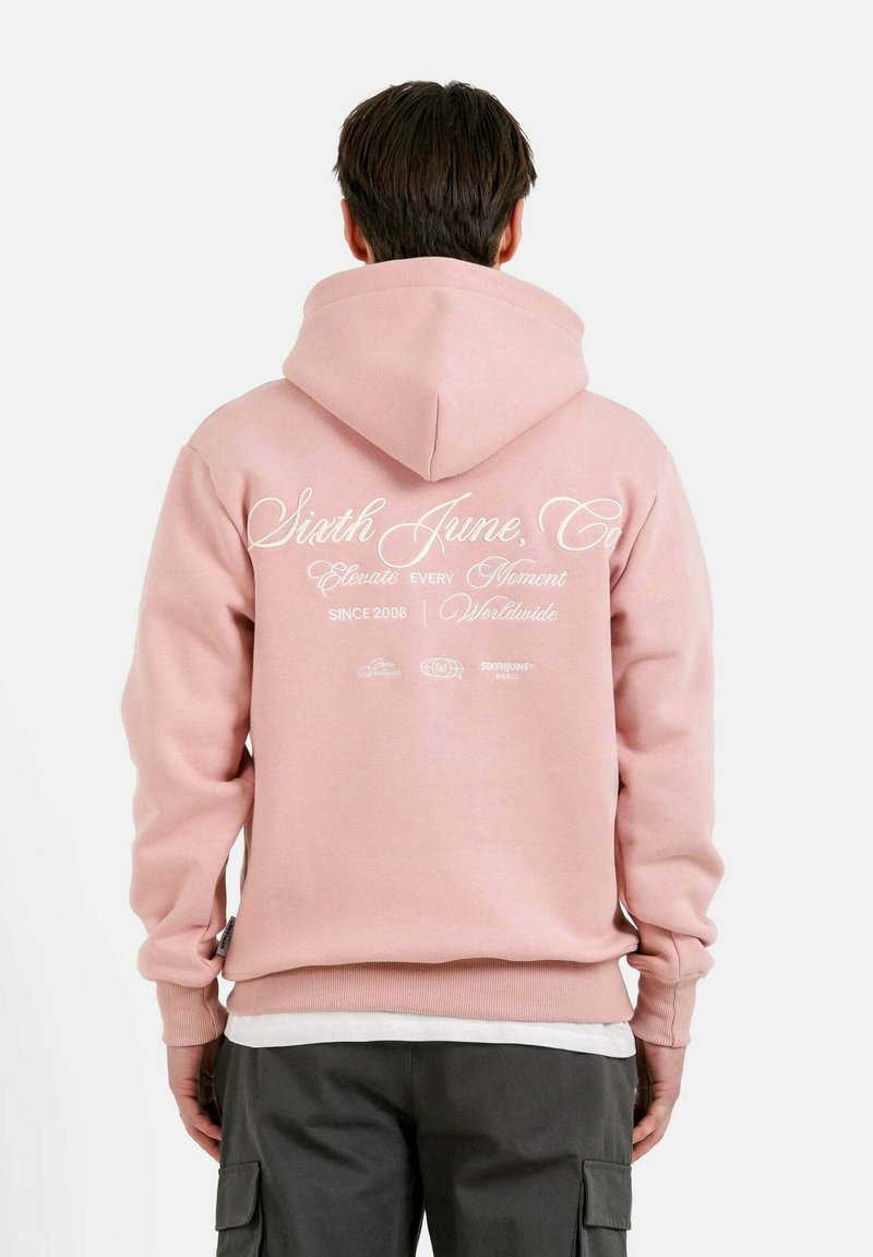 Sudadera con capucha rosa que presenta un gran texto de logo blanco en la parte posterior, puños y cinturilla acanalados. El material parece suave y cómodo.