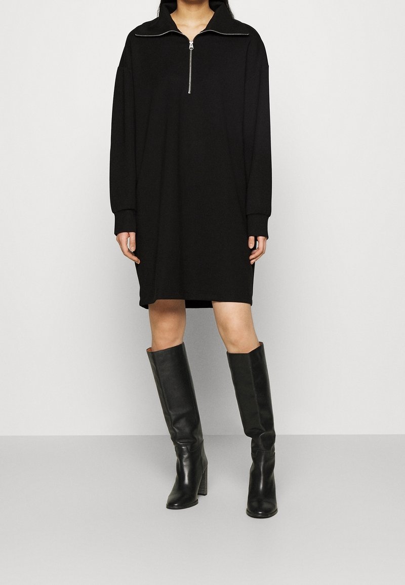 Svart oversized klänning med hög krage, halvdragkedja och långa ärmar, tillsammans med knähöga svarta läderstövlar.