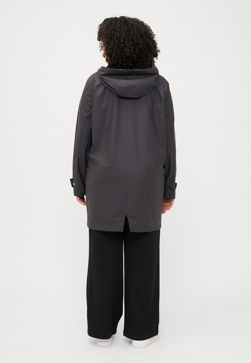 Een persoon met krullend haar draagt een lange, donkere jas met capuchon, zwarte wijde broek en witte schoenen, met de rug naar de camera tegen een neutrale achtergrond.