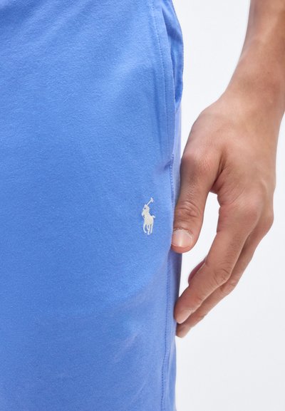 Χέρι ακουμπισμένο κοντά στην τσέπη ανοιχτό γαλάζιου παντελονιού με μικρό λευκό κεντητό λογότυπο παίκτη πόλο.