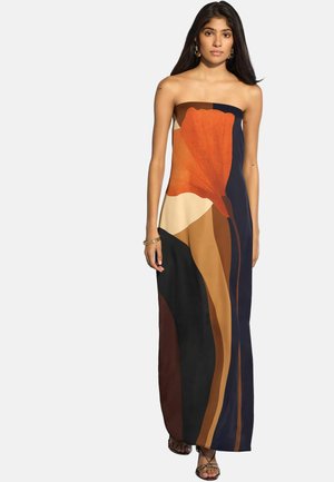 Kobieta o długich ciemnych włosach nosząca sukienkę maxi bez ramiączek z dużym abstrakcyjnym kwiatowym wzorem w kolorach pomarańczowym, brązowym, beżowym, czarnym i granatowym.