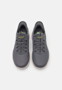 Grå träningsskor med meshinlägg, elastiska snören och gummisula; har gröna detaljer och Skechers-logotyp på tungan.