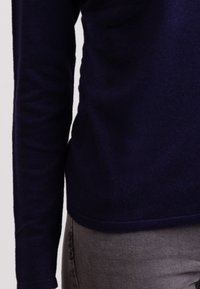 Námořnický svetr s dlouhým rukávem vyrobený z měkkého, texturovaného pleteného materiálu, s kulatým lemem a těsnými manžetami, v kombinaci se světle šedými kalhotami.