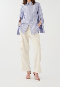 Chemise rayée bleue et blanche à boutons avec manches ouvertes et poche poitrine, portée avec un pantalon cargo beige et des talons nus à brides.