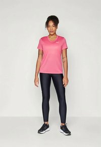 Moteris stovi vilkėdama rožinės spalvos trumpomis rankovėmis sportinį marškinėlį, juodas legginsus ir juodus bėgimo batus prieš paprastą foną.