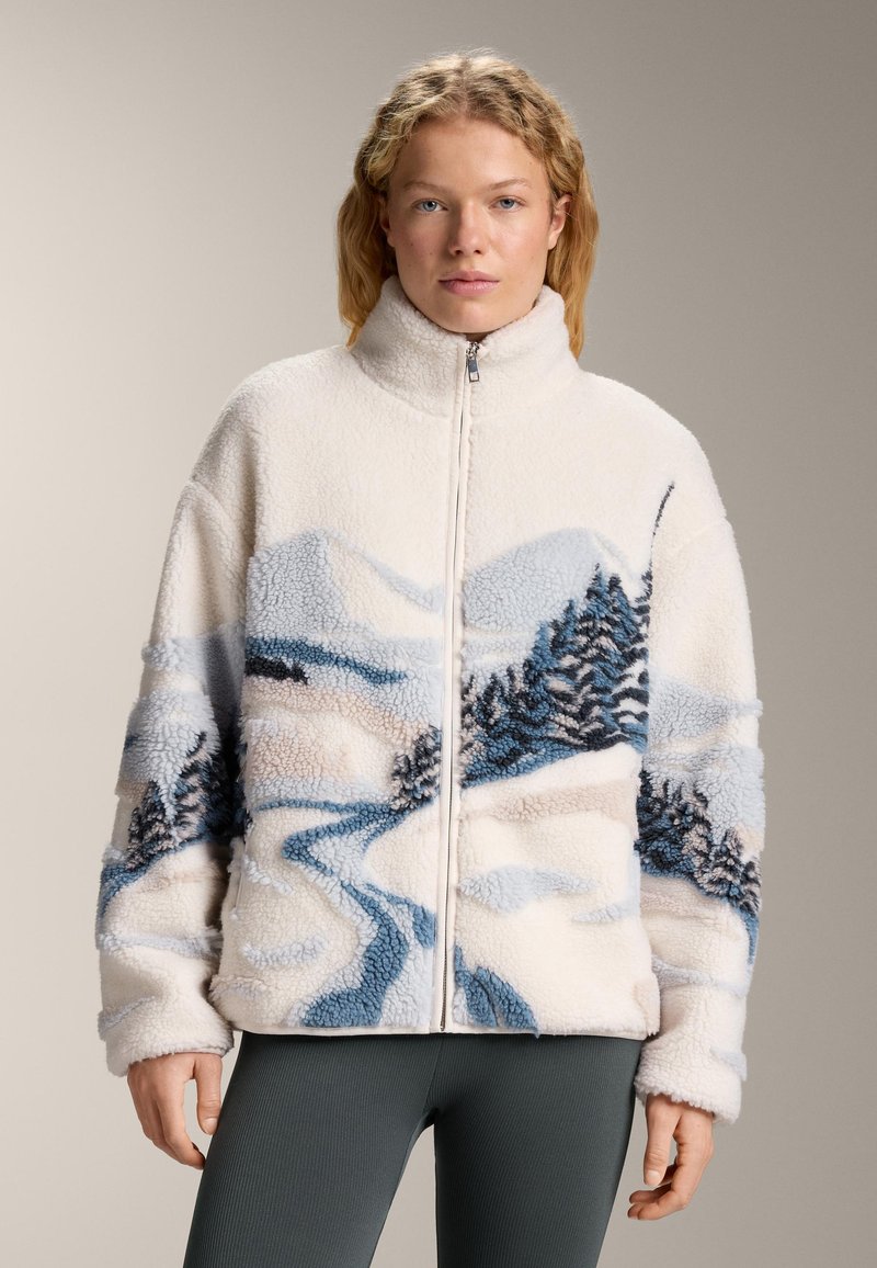 Ženska, oblečena v belo flis jakno z modrimi in črnimi vzorci gora ter dreves, stoji na enobarvni bež podlagi.