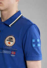 Giovane con indosso una polo blu con toppe, tra cui un logo circolare, del testo e una bandiera finlandese sulla manica, su uno sfondo grigio.