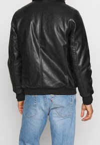 Czarna skórzana kurtka z prążkowanymi mankietami i dołem. Gładka faktura, lekko dopasowany fason, widok od tyłu. W pasie widoczne niebieskie dżinsy.