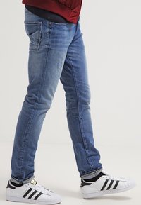 Jeans de denim azul ajustados con un ligero desgaste y un detalle sutil en el bolsillo trasero, combinados con zapatillas Adidas blancas y negras.