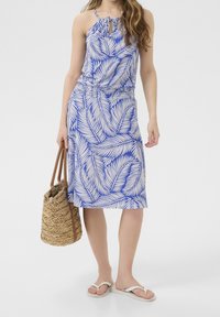 Robe à motif bleu et blanc avec un décolleté halter, taille cintrée et jupe à mi-jarret. Le mannequin tient un sac en osier. Tongs aux pieds.