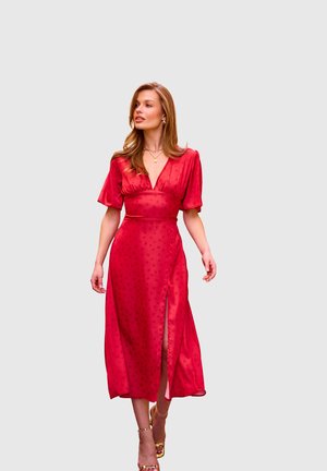 Kobieta pewnie idąca w czerwonej sukience o średniej długości z bufkami, dekoltem w serek i rozcięciem po boku, nosząca złote szpilki i biżuterię.