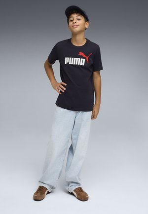 Niño con camiseta azul marino de Puma, vaqueros anchos azul claro y zapatillas marrones, de pie con la mano en la cadera contra un fondo gris.