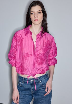 Mujer con cabello largo y oscuro, vistiendo una blusa bordada de color rosa brillante atada a la cintura y jeans de mezclilla azul, de pie frente a un fondo liso.