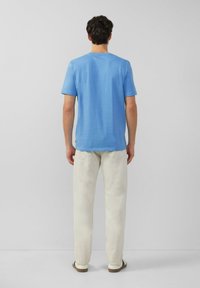 Man die met zijn rug naar de camera staat, gekleed in een blauwe korte mouw shirt, lichte beige broek en witte instapschoenen, tegen een effen achtergrond.