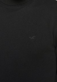 Zwarte gebreide trui met een hoge halslijn en een discreet geborduurd logo op de borst. Gladde textuur, slim fit ontwerp en geen zichtbare patronen.