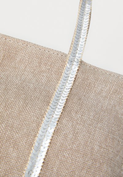 Sac en tissu beige avec une surface texturée, présentant une bordure en sequins argentés brillants le long de la anse pour un détail décoratif.