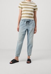 Person som bär en beige- och vit randig kortärmad skjorta, ljusblå joggerjeans med dragsko och svarta öppna sandaler med stort spänne.