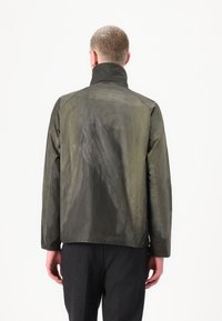 Casaco impermeável caqui com colar alto, textura suave e design minimalista. Apresenta aberturas laterais e um ajuste elegante.