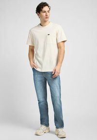 Cream cotton t-shirt with a chest pocket, paired with light blue denim jeans. The outfit has a relaxed fit and casual design.