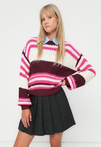 Suéter de malha com listras rosa, branca e maroon, apresentando uma borda canelada e mangas largas, combinado com uma saia plissada preta.