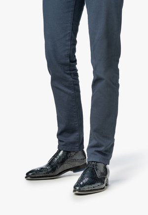 Botas de tobillo de cuero negro con un patrón texturizado similar a reptiles. Combinadas con pantalones ajustados azul oscuro, que exhiben un diseño elegante.