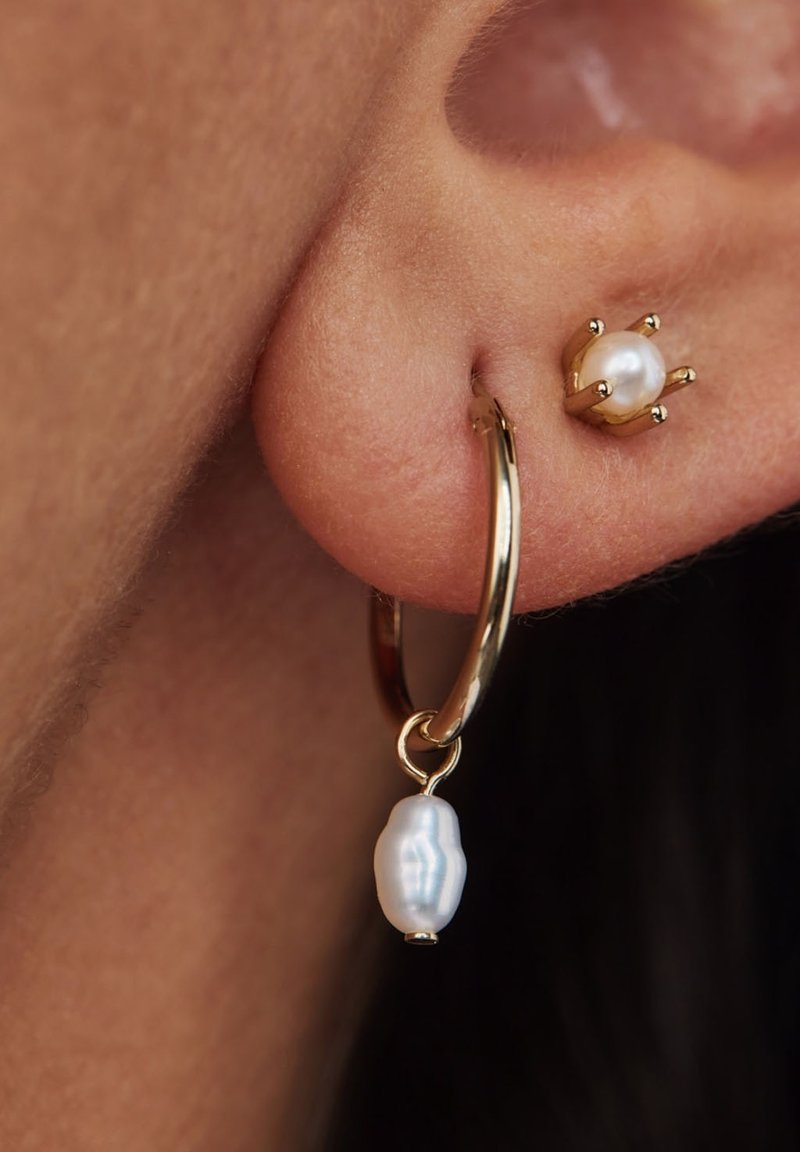 Goldene Creolen mit einer hängenden, strukturierten weißen Perle und einem kleinen Perlenstecker. Nahaufnahme am Ohrläppchen, die Details und das Finish hervorhebt.