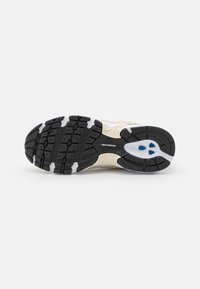 Semelle d'une basket New Balance beige avec une bande de roulement en caoutchouc noir, des accents blancs et des éléments de coussinage bleus, présentée sur un fond blanc.