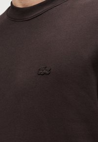 Sweat-shirt marron foncé avec un col rond côtelé, arborant un logo noir brodé discret sur la poitrine, fabriqué en tissu doux mélangé en coton.
