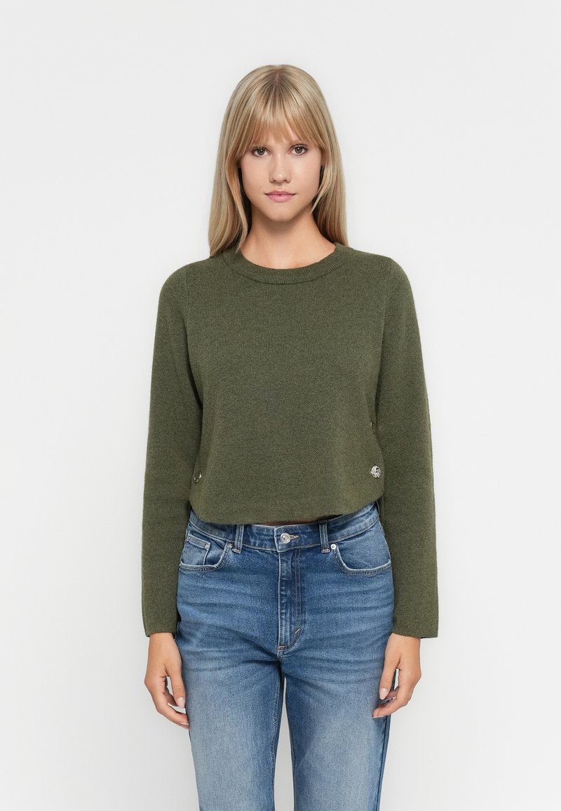 Maglione corto a maniche lunghe in tessuto a maglia verde oliva con scollatura rotonda e bottoni decorativi sui lati. Abbinato a jeans blu.