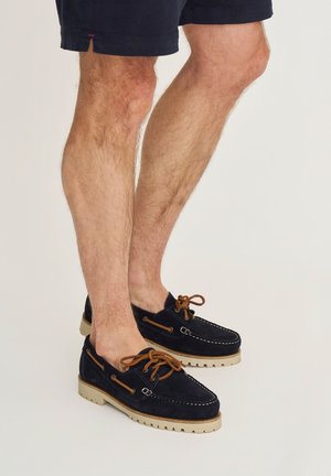 Man met marineblauwe suède bootsschoenen met bruine veters en beige zolen, gecombineerd met een donkere korte broek, staand op een lichte achtergrond.