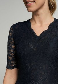 Dunkelblaues Spitzenoberteil mit kurzen Ärmeln, über einem gewellten V-Ausschnitt und floralen Mustern im gesamten Stoff. Strukturierte Optik.