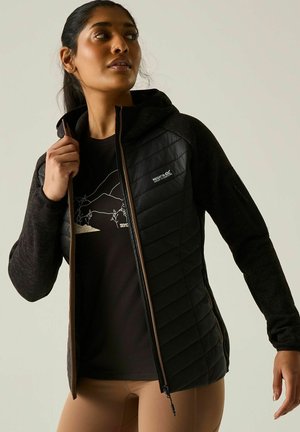 Mujer con chaqueta negra acolchada con capucha y cremallera frontal, superpuesta a una camiseta negra con gráfico, posando sobre un fondo liso.