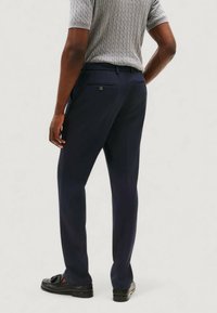 Pantaloni blu navy con una vestibilità sartoriale e una texture liscia. Presentano una tasca posteriore con dettaglio a bottone. Abbinati a un top grigio a coste.