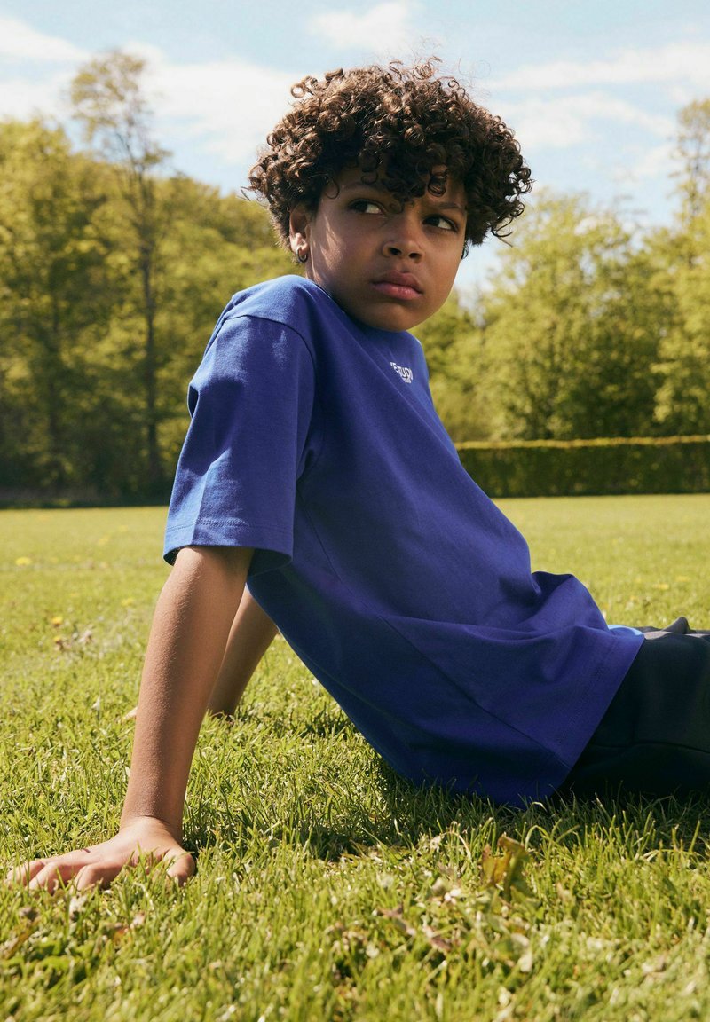 Enfant aux cheveux bouclés portant une chemise bleue assis sur l'herbe, s'appuyant sur les mains en arrière, regardant sur le côté avec des arbres et le ciel en arrière-plan.