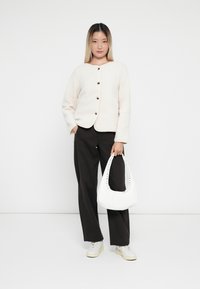 Gilet en crème flou avec col rond et boutons marron, associé à un pantalon noir ample et un sac blanc avec des clous. Baskets blanches.