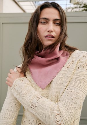 Mladá žena s dlouhými hnědými vlasy, která má na sobě krémový pletený svetr a purpurovou šátek kolem krku, stojí venku před šedým pozadím.