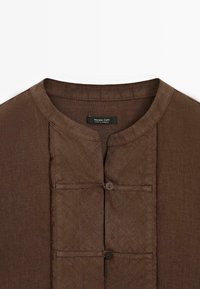 Camicia in lino marrone con scollo rotondo, chiusura frontale con bottoni e design minimalista. Caratterizzata da cuciture sottili e finitura strutturata.