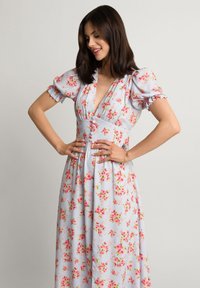 Kwiatowa sukienka maxi na jasnoniebieskim tle, z bufiastymi rękawami, głębokim dekoltem i detalami w postaci guzików w talii. Charakteryzuje się zwiewną sylwetką.