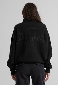 Svart oversized hettegenser med høy krage. Har hevet tekst og et bladdesign på baksiden. Laget av et mykt, teksturert stoff.