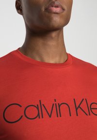 T-shirt rossa in cotone caratterizzata da un colletto rotondo e maniche corte, con "Calvin Klein" stampato in grandi lettere nere sul petto.
