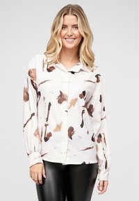 Langermet bluse i kremfarge med abstrakte brune og brune mønstre, med knappelukking foran og en avslappet passform, kombinert med svarte leggings.